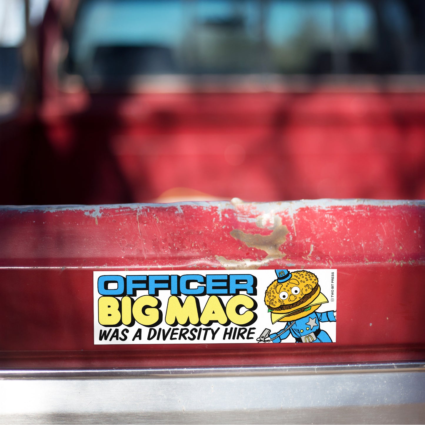 A vinyl bumper magnet reading "OFFICER BIG MAC WAS A DIVERSITY HIRE" on red truck tailgate.