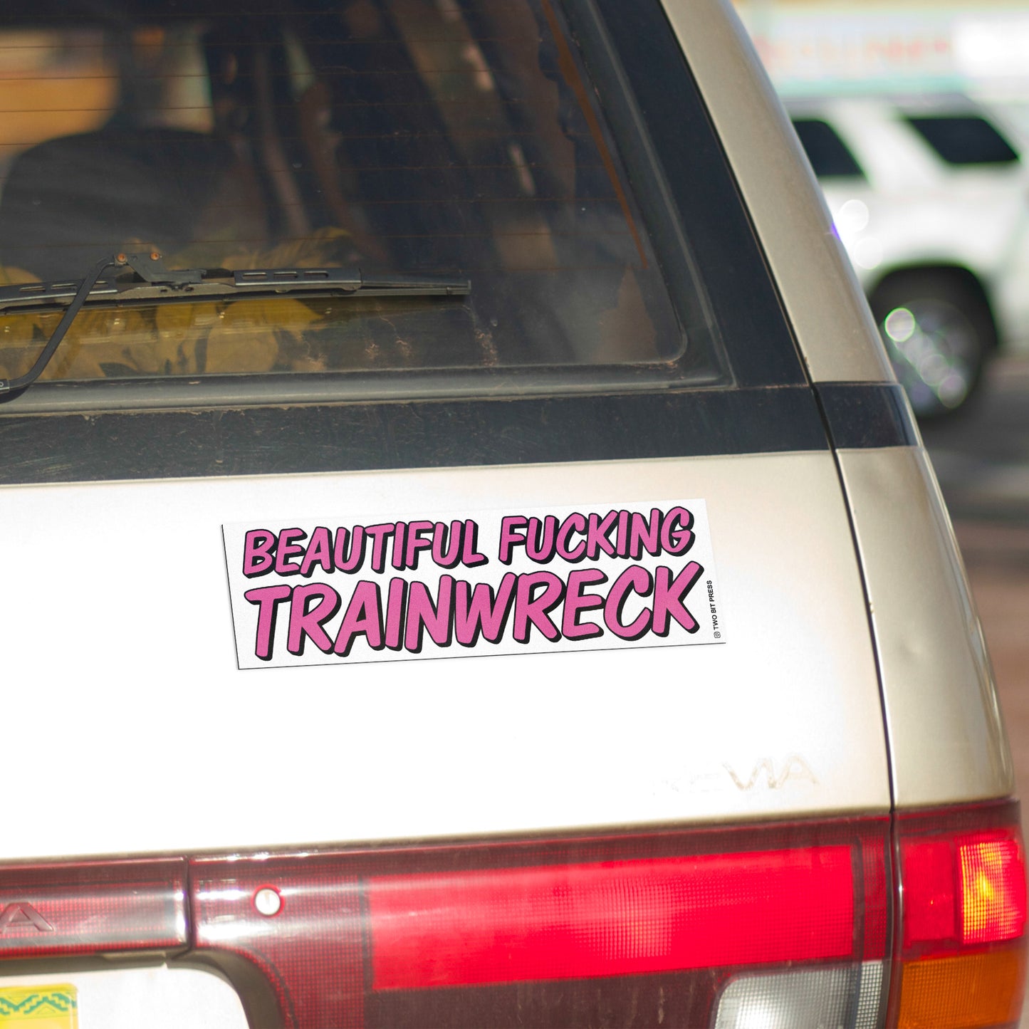 A vinyl bumper magnet reading "BEAUTIFUL FUCKING TRAINWRECK" on gold minivan.
