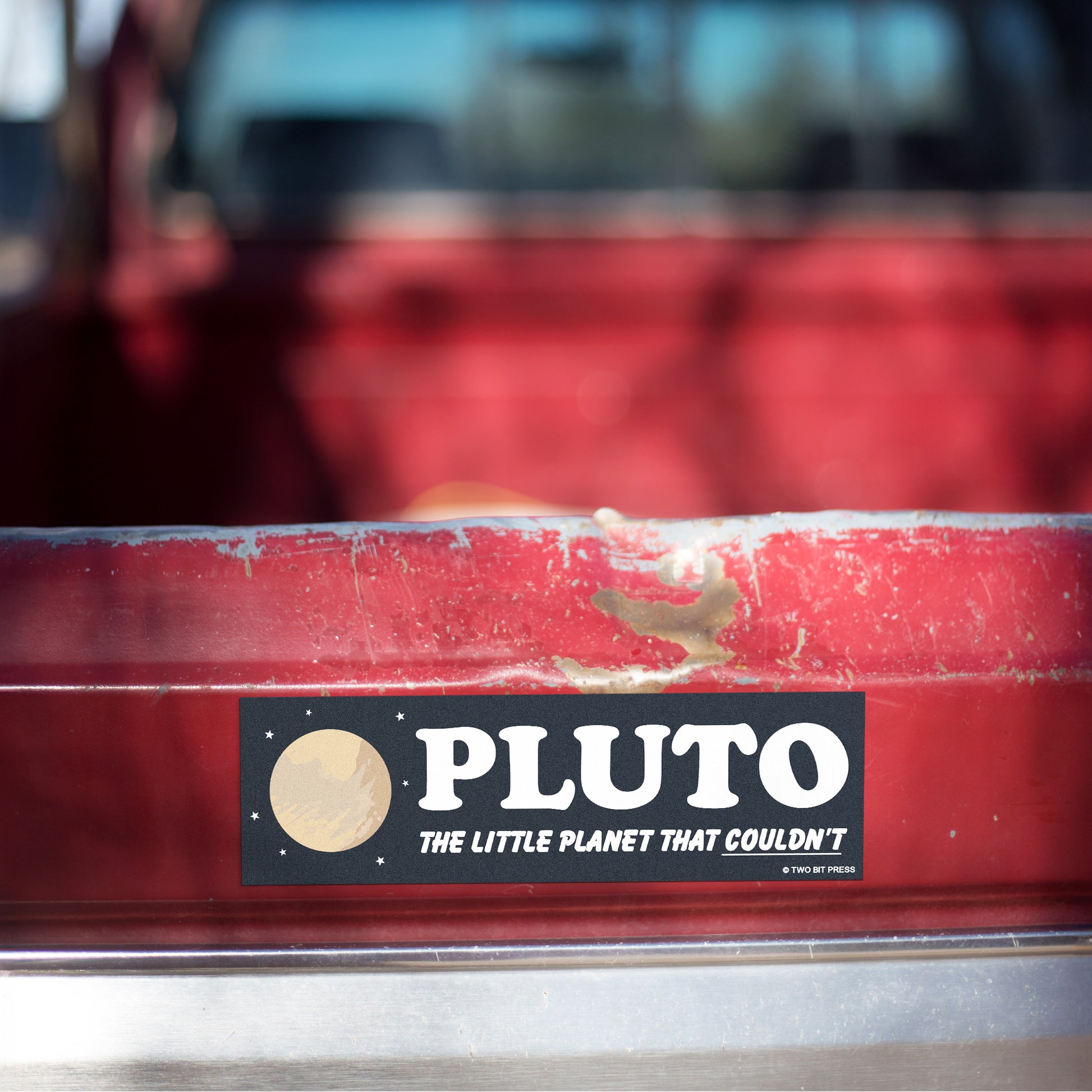 A vinyl bumper magnet showing Pluto and the text "PLUTO Little Planet" on red truck tailgate.