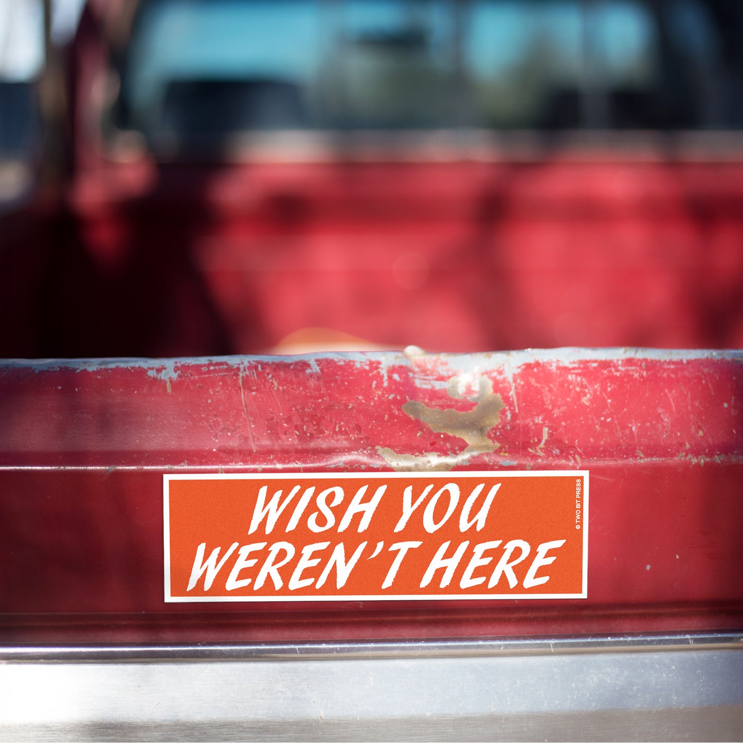 A vinyl bumper magnet reading "WISH YOU WEREN'T HERE" on red truck tailgate.