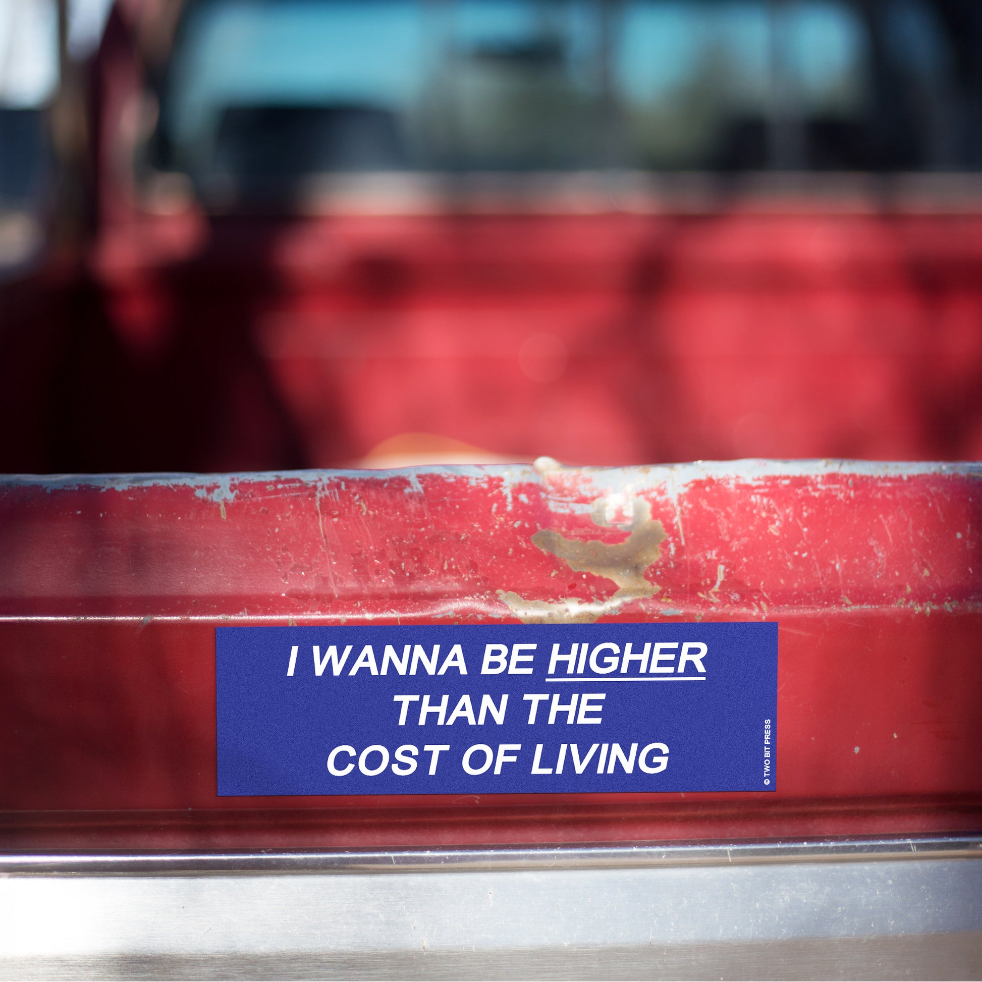 A vinyl bumper magnet reading "I WANNA BE HIGHER THAN THE COST OF LIVING" on red truck tailgate.