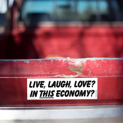 A vinyl bumper magnet reading "LIVE, LAUGH, LOVE? IN THIS ECONOMY?" on red truck tailgate.