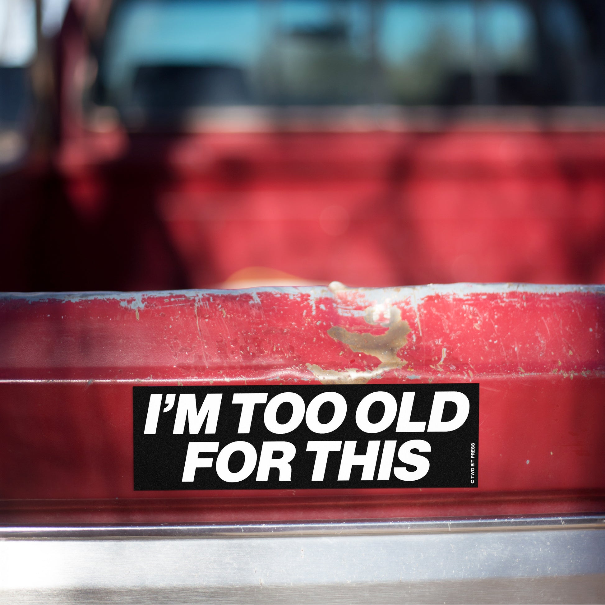 A vinyl bumper magnet reading "I'M TOO OLD FOR THIS" on red truck tailgate.
