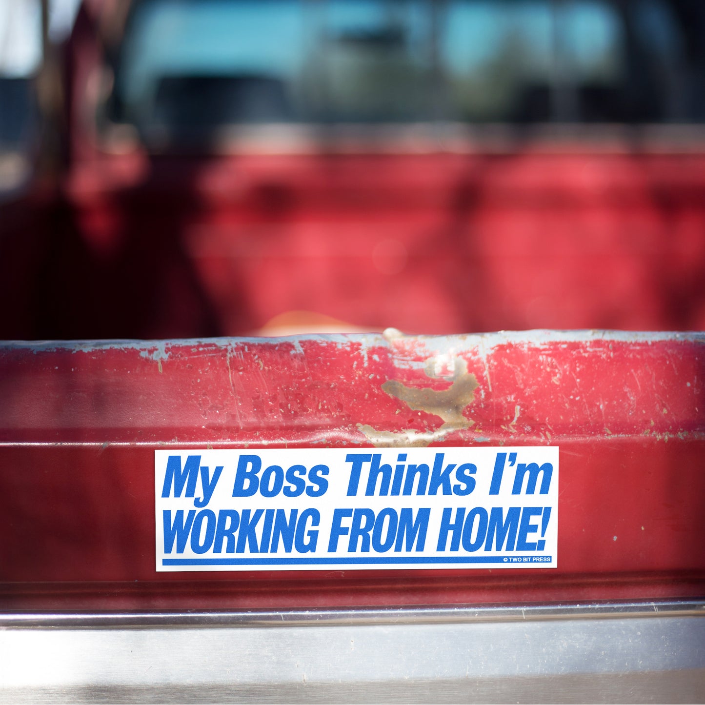 A vinyl bumper magnet reading "My Boss Thinks I'm WORKING FROM HOME" on red truck tailgate.