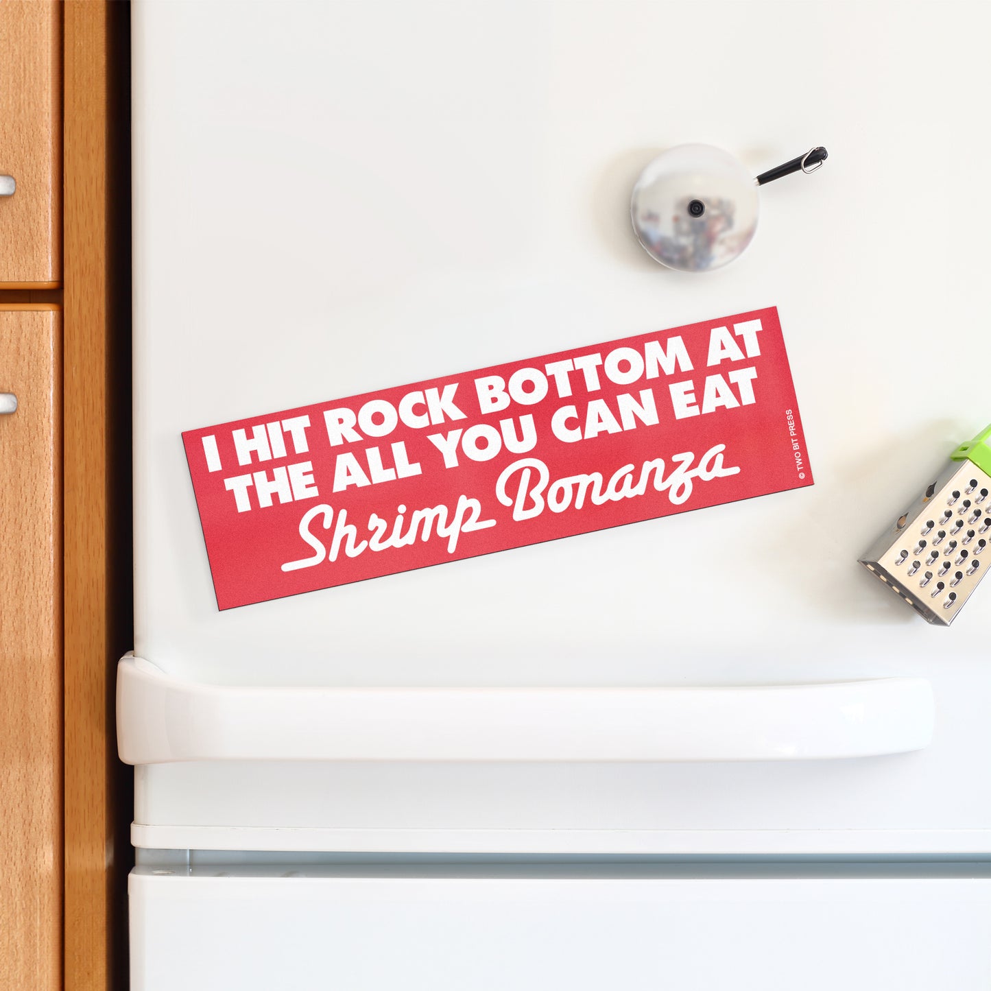 A vinyl bumper magnet reading "I HIT ROCK BOTTOM AT THE ALL YOU CAN EAT Shrimp Bonanza" on a white fridge.
