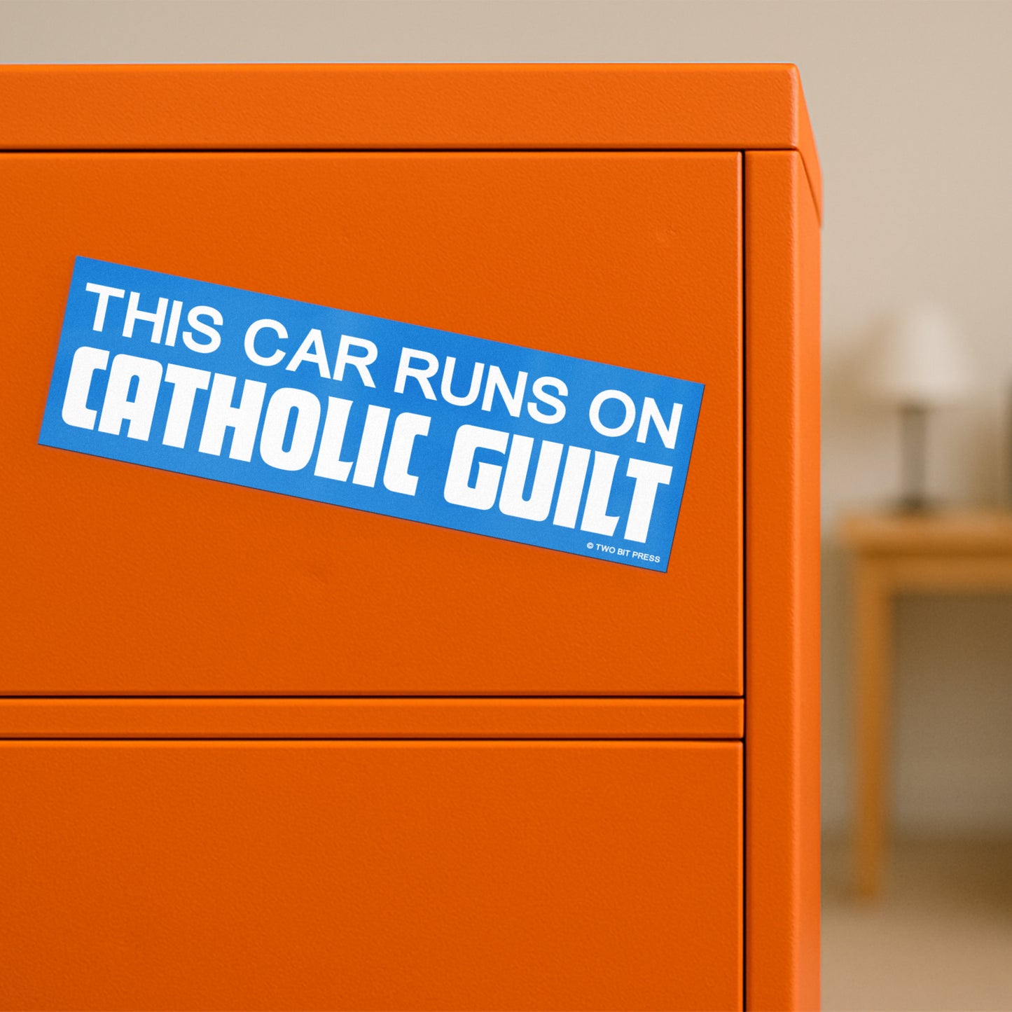 A vinyl bumper magnet reading "THIS CAR RUNS ON CATHOLIC GUILT" on orange filing cabinet.