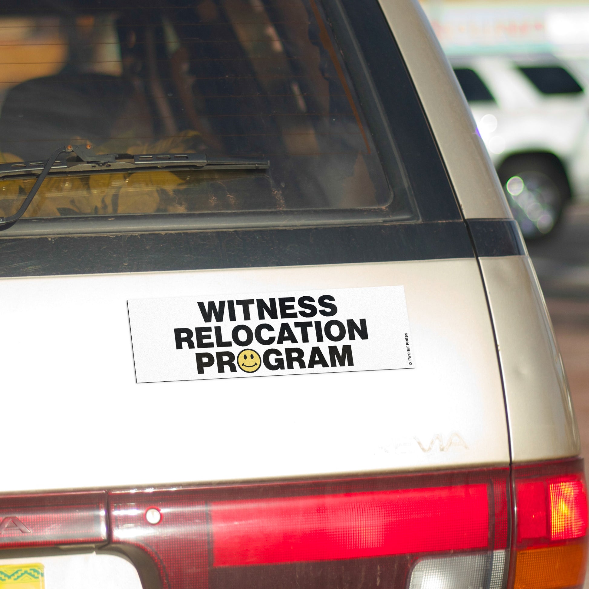 A vinyl bumper magnet reading "WITNESS RELOCATION PROGRAM" on gold minivan.