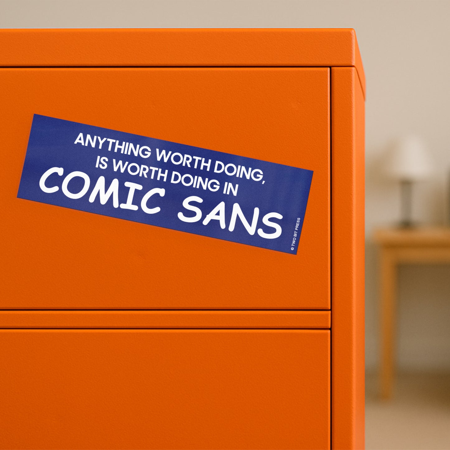 A vinyl bumper magnet reading "ANYTHING WORTH DOING IS WORTH DOING IN COMIC SANS" on orange filing cabinet.