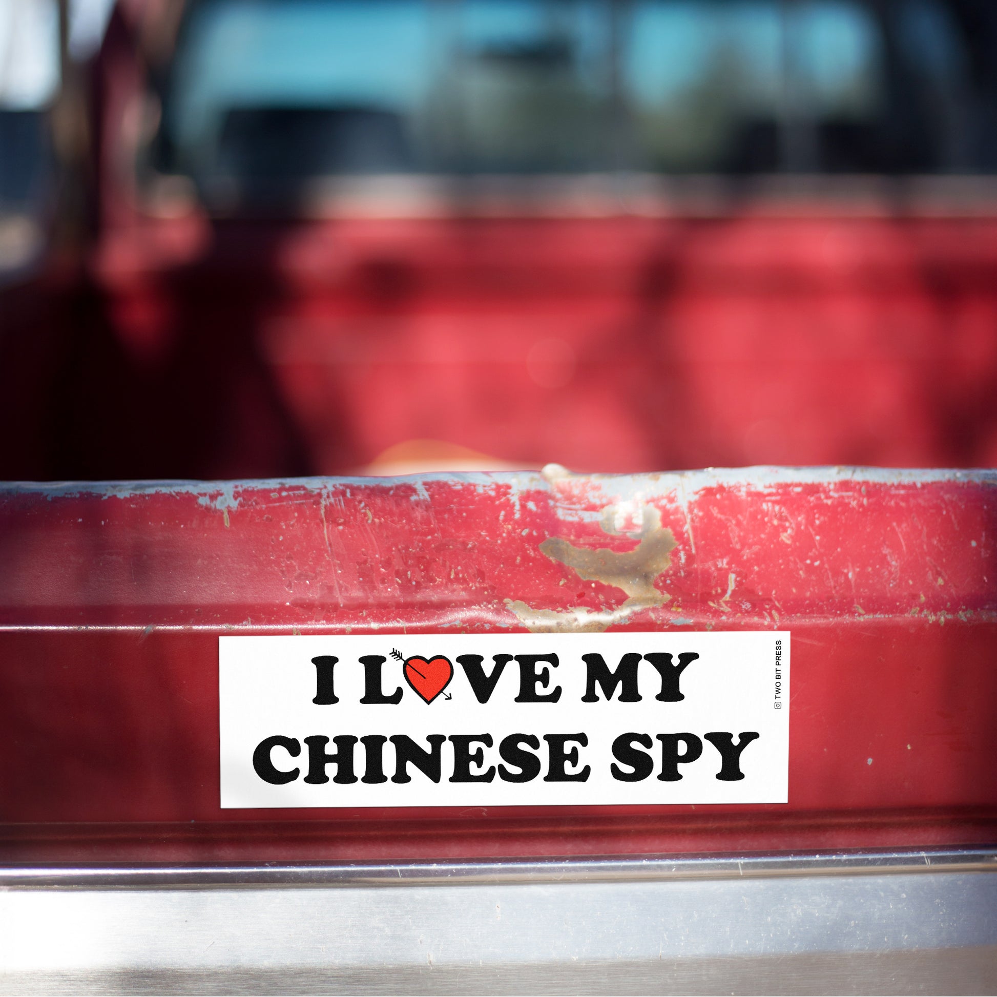 A vinyl bumper magnet reading "I LOVE MY CHINESE SPY" on red truck tailgate.