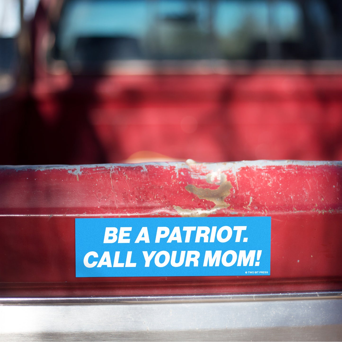 A vinyl bumper magnet reading "BE A PATRIOT. CALL YOUR MOM!" on red truck tailgate.