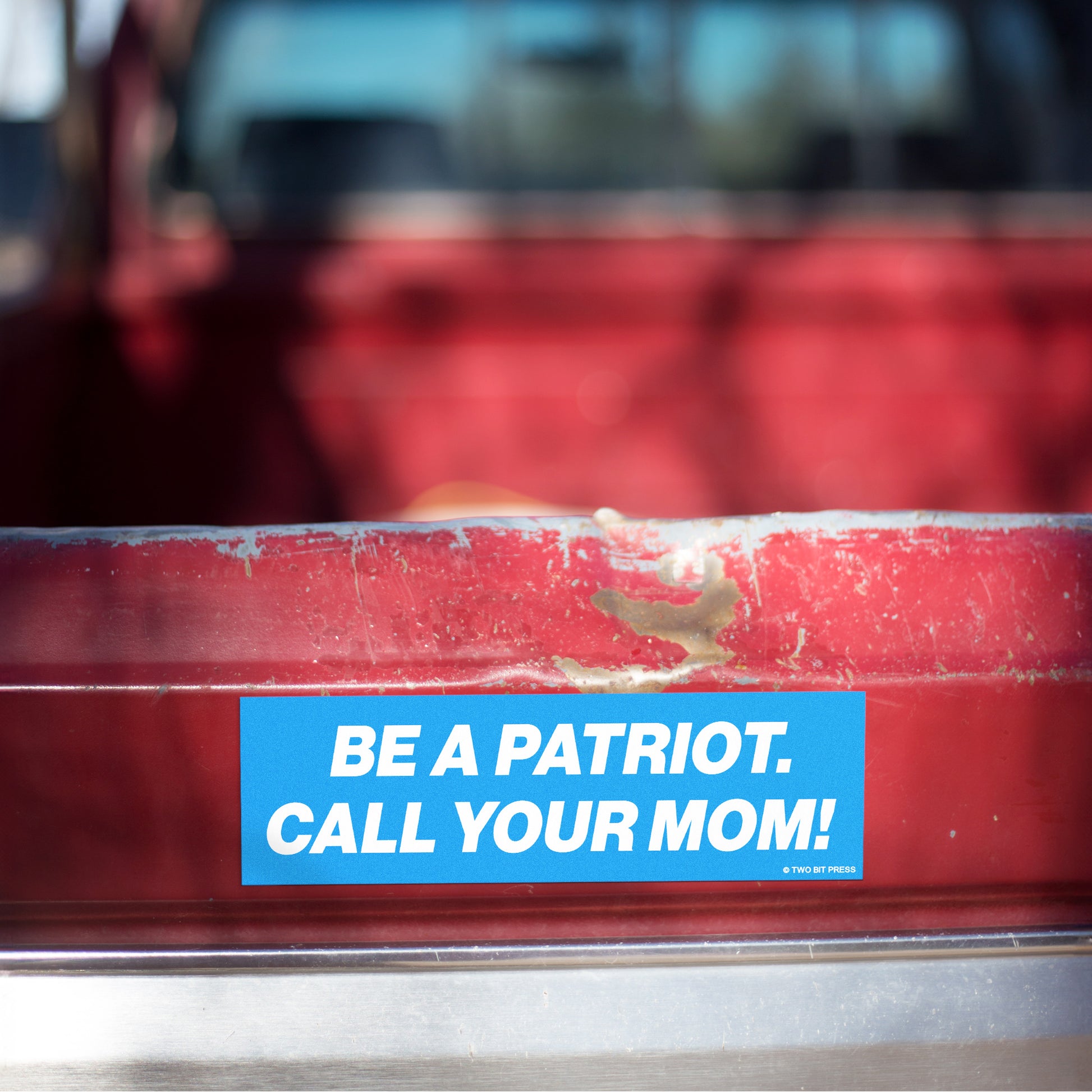 A vinyl bumper magnet reading "BE A PATRIOT. CALL YOUR MOM!" on red truck tailgate.