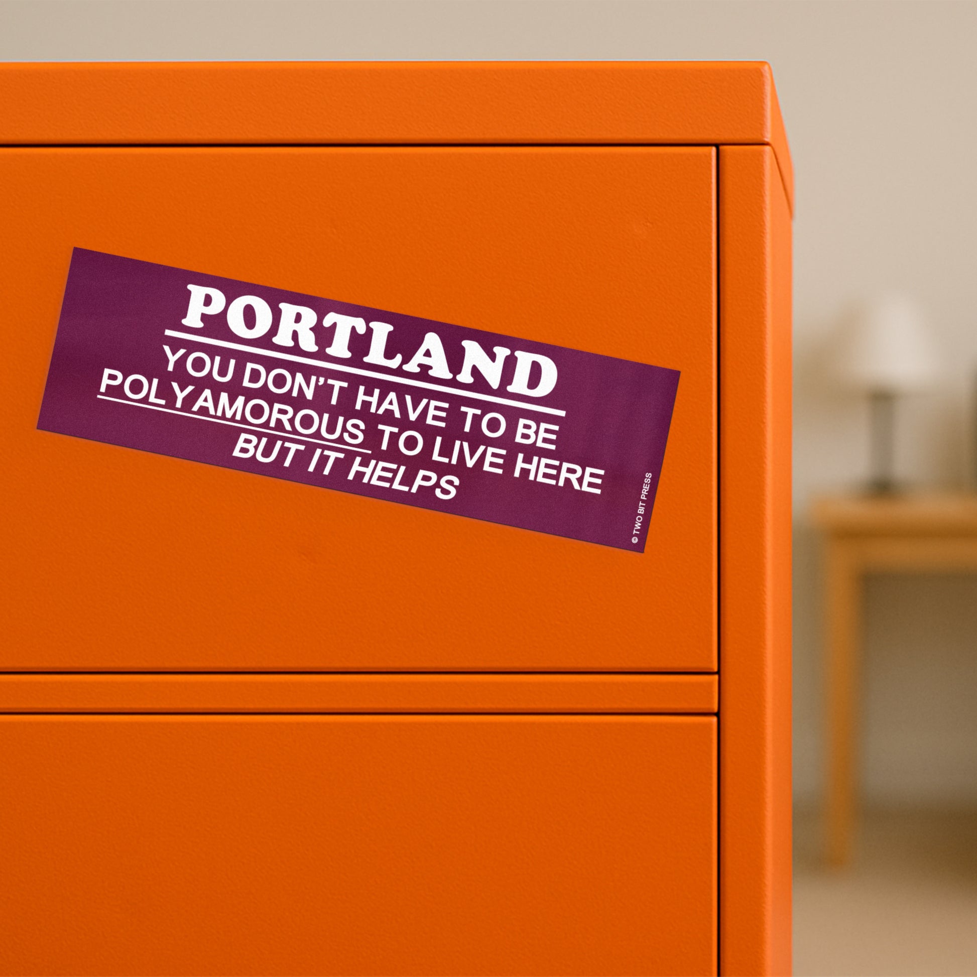 A vinyl bumper magnet reading "PORTLAND YOU DON'T HAVE TO BE POLYAMOROUSTO LIVE HERE BUT IT HELPS" on orange filing cabinet.