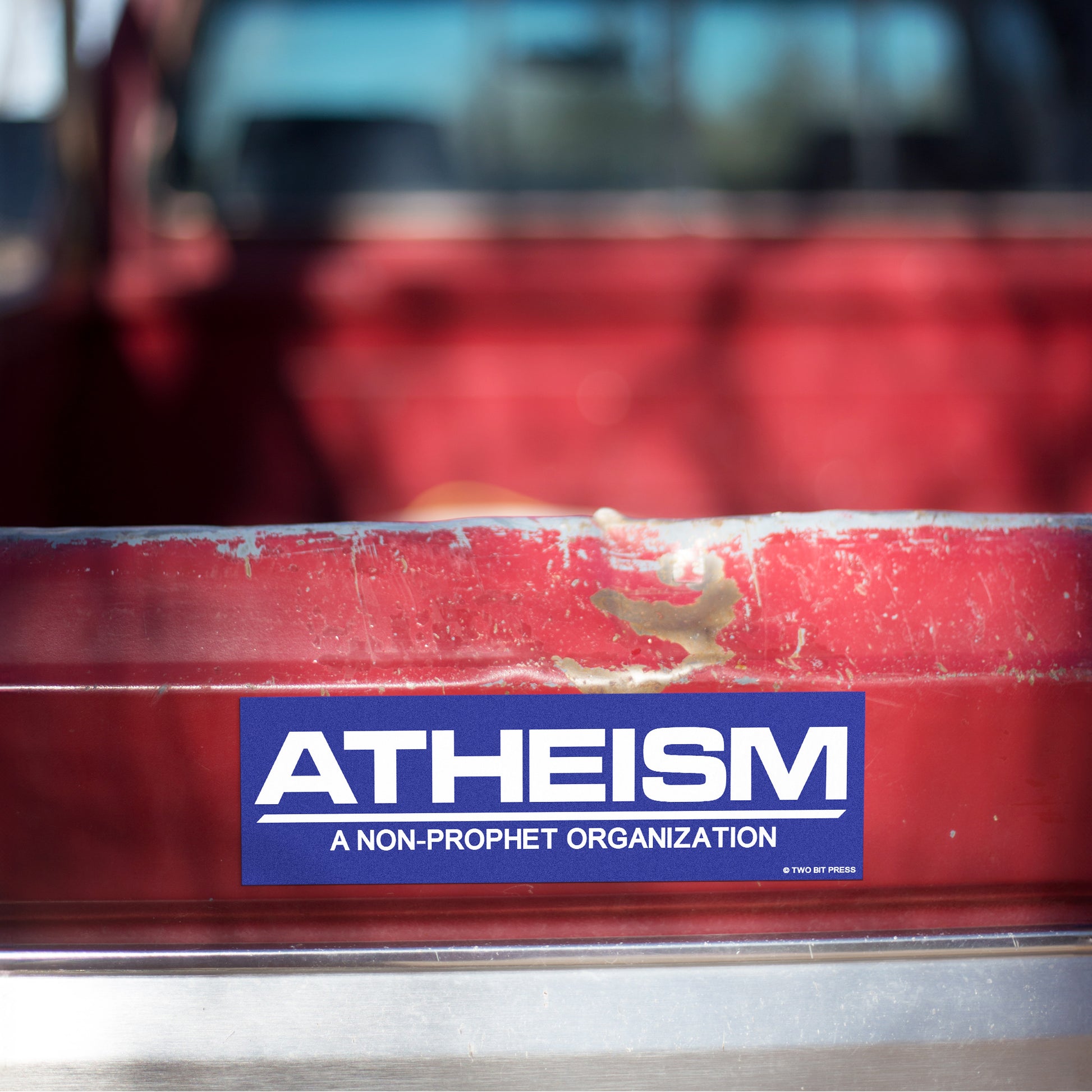 A vinyl bumper magnet reading "ATHEISM A NON-PROPHET ORGANIZATION" on red truck tailgate.