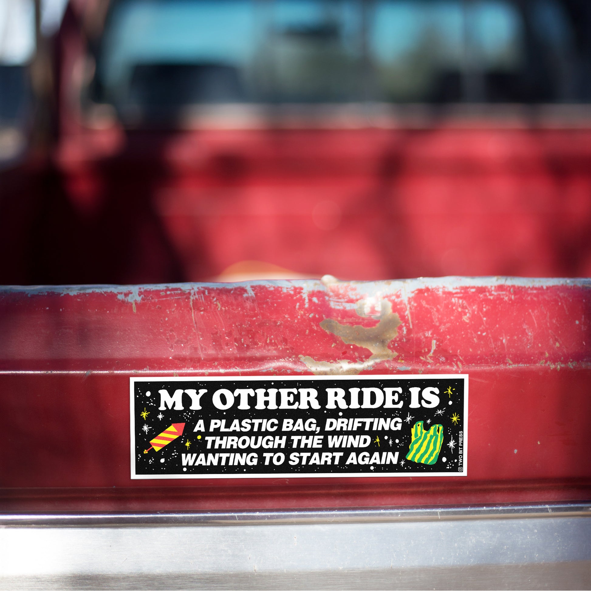 A bumper magnet reading "MY OTHER RIDE IS A PLASTIC BAG, DRIFTING THROUGH THE WIND WANTING TO START AGAIN" on red truck tailgate.