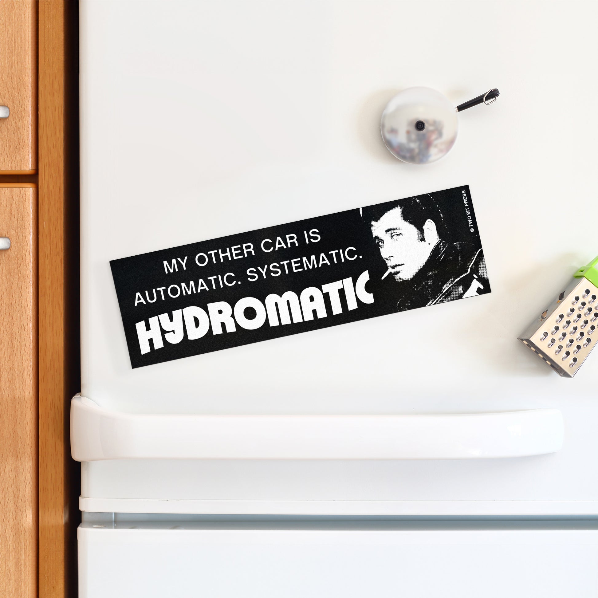 A bumper magnet showing John Travolta and the text "MY OTHER CAR IS AUTOMATIC SYSTEMATIC HYDROMATIC" on a white fridge.