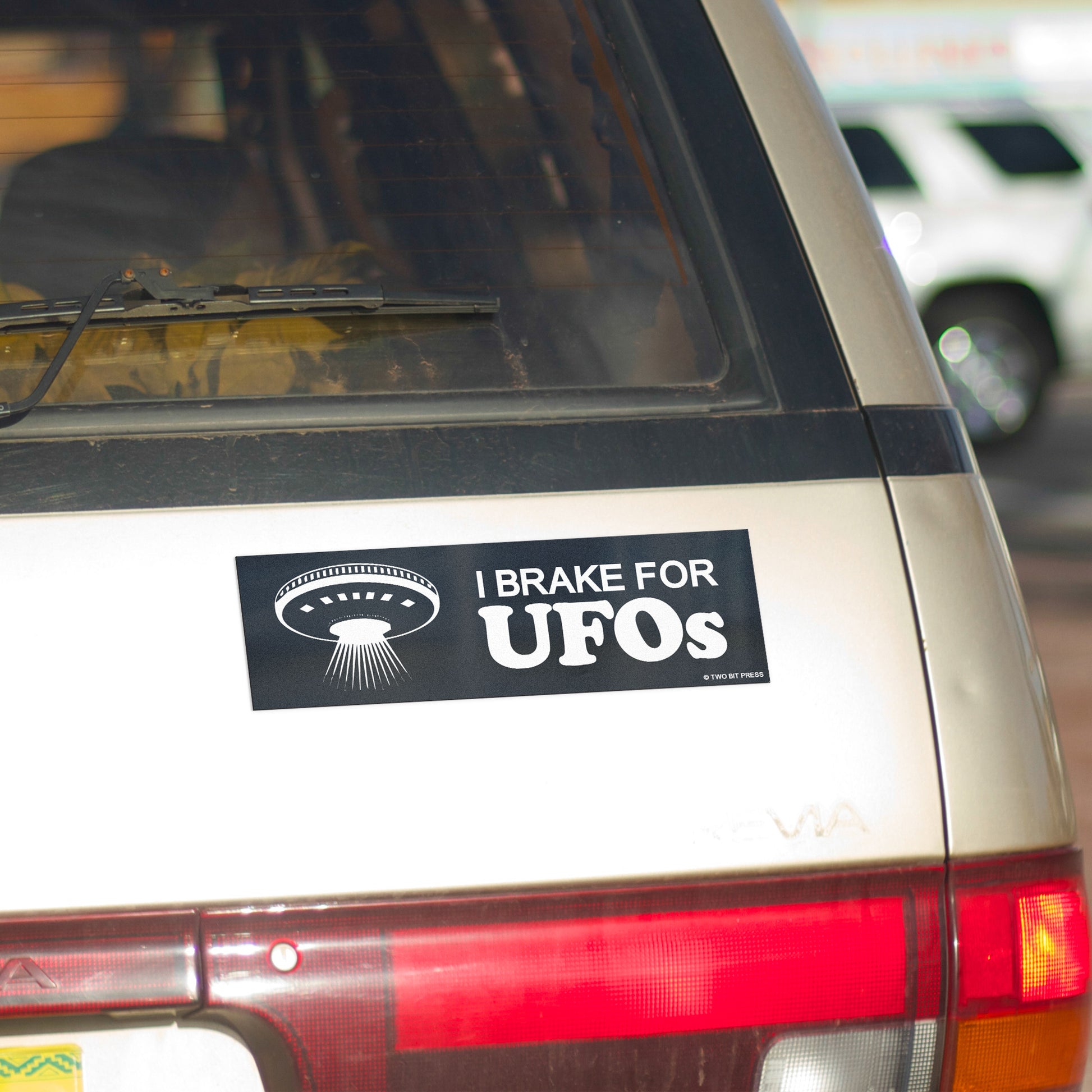 A vinyl bumper magnet showing a flying saucer and the text "I BRAKE FOR UFOS" on gold minivan.