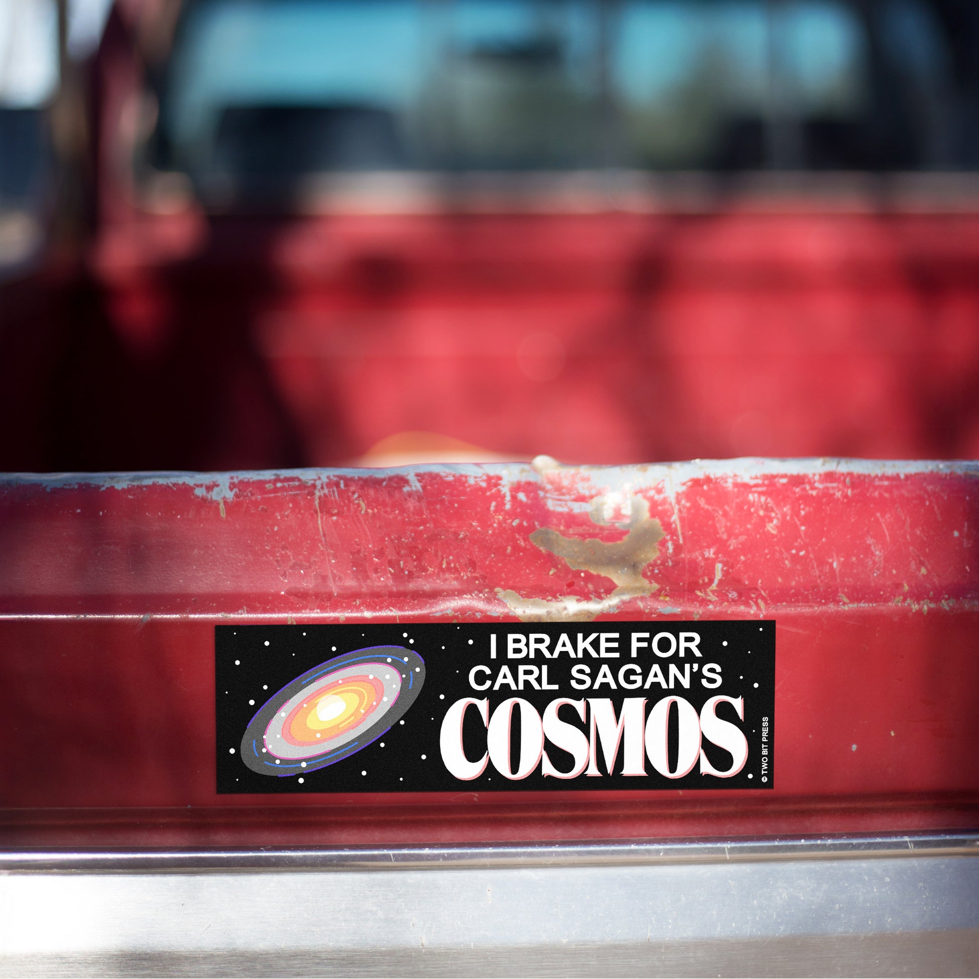 A vinyl bumper magnet showing a galaxy graphic and the text "I BRAKE FOR CARL SAGAN'S COSMOS" on red truck tailgate.