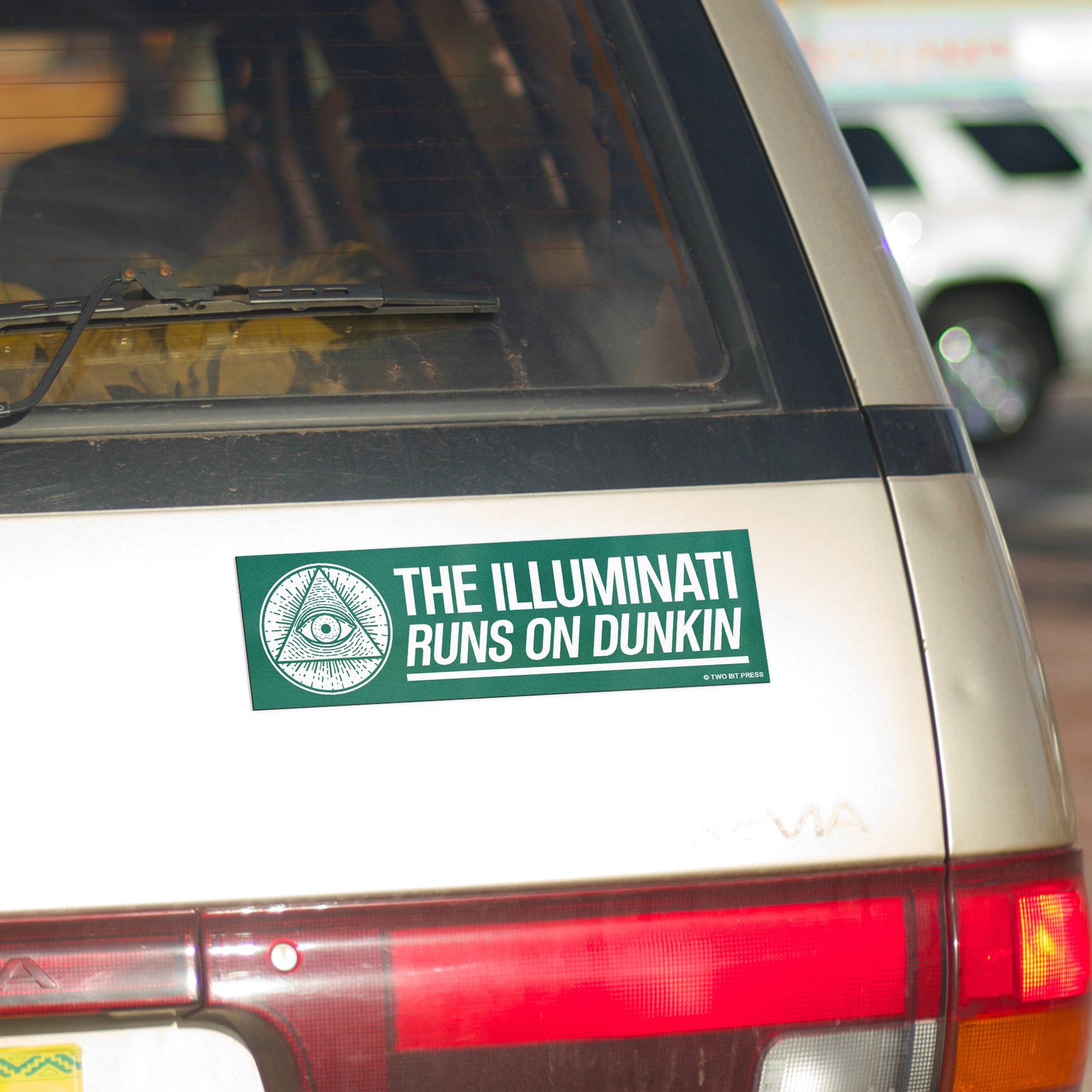 A vinyl bumper magnet reading "THE ILLUMINATI RUNS ON DUNKIN" on gold minivan.