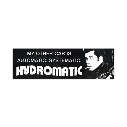 A bumper magnet showing John Travolta and the text "MY OTHER CAR IS AUTOMATIC SYSTEMATIC HYDROMATIC" on a white background.